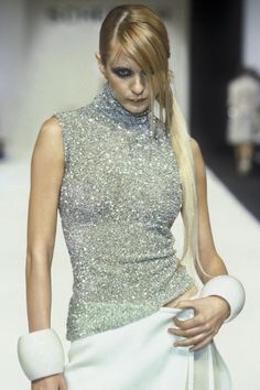 a woman with long blonde hair walking down a runway wearing a silver top and white skirt