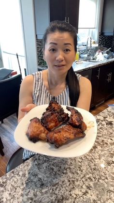 a woman sitting at a kitchen table holding a plate with meat on it and smiling