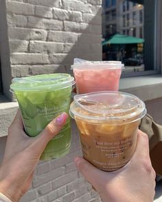 two people holding up drinks in front of a brick building on a sunny day, one is green and the other is pink