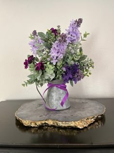 a vase with purple flowers and greenery in it on top of a piece of wood