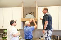 three men standing in a kitchen looking at something on the cabinet above them and pointing to it