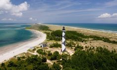 an aerial view of a lighthouse on the beach with trees and water in the background