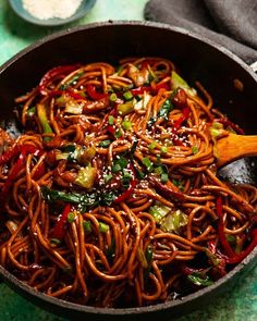 a wok filled with noodles and vegetables on top of a green tablecloth next to a wooden spoon