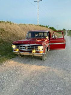 an old red pick up truck parked on the side of a road next to a tall grass covered hill