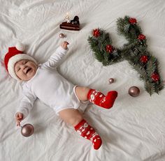 a baby laying on top of a white bed next to christmas decorations and baubles