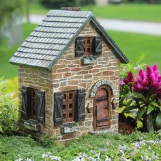 a small brick house sitting on top of a lush green field next to purple flowers