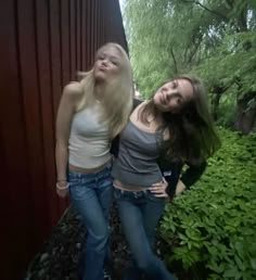 two young women standing next to each other in front of a red wall and trees