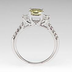 This beautiful yellowish green sapphire ring is centered with one (1) cut-cornered square mixed cut natural sapphire weighing 1.33 carats and set into a four-prong head. The sapphire is flanked to each side by one (1), channel/prong set, square modified brilliant cut diamond. The top face of the shank is accented with five (5), prong set, round brilliant cut diamonds. The ring measures 6.6mm at the top, rises 7.3mm above the finger, tapering to 1.6mm wide and 1.3mm thick at the base of the shank. The ring is currently size 5.5. Yellow Emerald Cut Diamond Ring, Gia Certified Asscher Cut Sapphire Ring, Gia Certified Square Cut Sapphire Diamond Ring, Square Cut Sapphire Ring With Diamond, Green Sapphire Ring, Yellowish Green, Three Stone Engagement Ring, Stone Engagement Ring, Three Stone Engagement