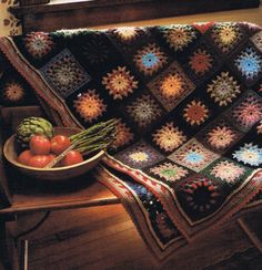 a crocheted blanket sitting on top of a wooden bench next to a bowl of fruit and vegetables