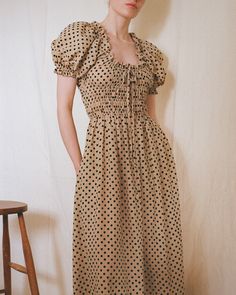 a woman in a polka dot dress standing next to a wooden stool and white wall