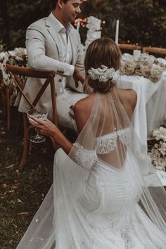 a bride and groom sitting at a table with flowers in their hair, holding wine glasses