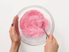 two hands holding a spoon over a pink substance in a glass bowl on a white surface