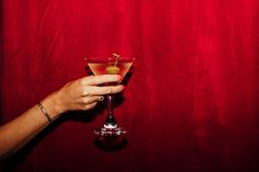 a hand holding a martini glass with an olive garnish on the rim, against a red background