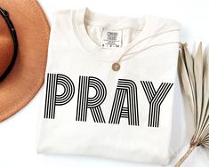 a white shirt with the word pray on it next to a hat and feather quill