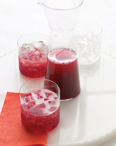 three glasses filled with red liquid on top of a white tablecloth next to ice cubes