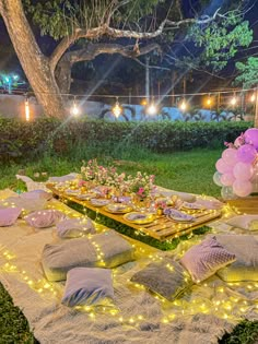 a table set up for a party with pink and purple balloons on it, surrounded by lights