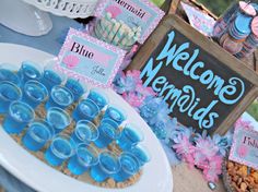 a table topped with lots of desserts next to a chalkboard sign that says welcome mermaids