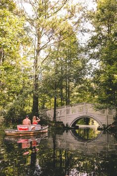 two people in a small boat on the water near a bridge and trees with leaves