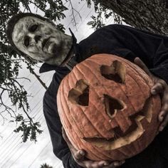 a man holding a carved pumpkin in front of a tree with a creepy face on it