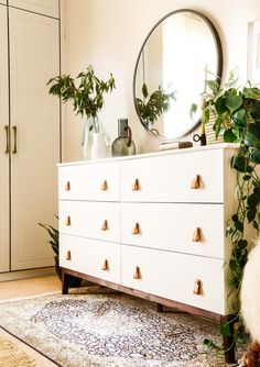 a white dresser topped with lots of green plants next to a tall mirror and potted plant