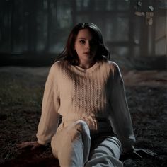 a woman sitting on the ground with her legs crossed, wearing white pants and a sweater