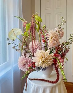 a white vase filled with lots of flowers on top of a wooden table next to a window