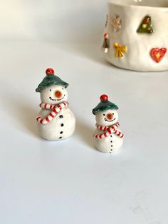 two small snowmen sitting next to each other on a white surface with a cup in the background