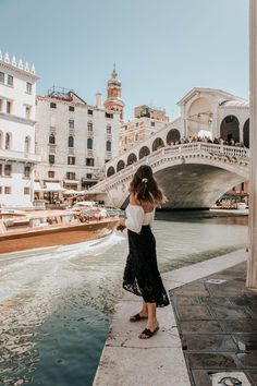 a woman standing on the side of a river next to a bridge