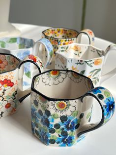 several colorful cups are lined up on a white tablecloth with flowers painted on them