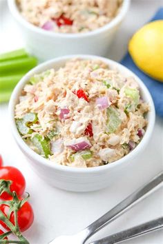 two bowls filled with tuna salad on top of a white table next to lemons and tomatoes