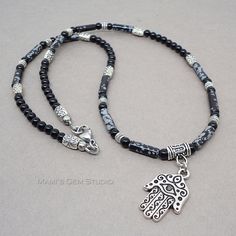 "This distinctive necklace features a metal pendant of Hamsa, known as an amulet shaped like a hand and is thought to protect against the evil eye. This detailed Hamsa charm is a made-in-USA high quality TierraCast product, which is made of pewter in antiqued fine silver finish. It hangs from a necklace that I designed with natural Snowflake Obsidian stone tube beads in black with whitish gray patches resembling snowflakes, and Black Onyx gemstone round beads. I have also added antiqued silver f Spiritual Silver Obsidian Necklace, Black Hand-strung Amulet Necklace, Black Amulet Style Beaded Necklaces, Black Amulet Necklace, Black Hand-strung Amulet Jewelry, Black Amulet Beaded Necklaces For Jewelry Making, Hand Of Hamsa, Mens Beaded Necklaces, Amulet Charm