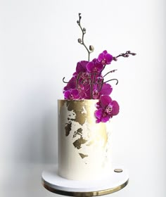 a white and gold cake with purple flowers on top is sitting on a stand against a wall