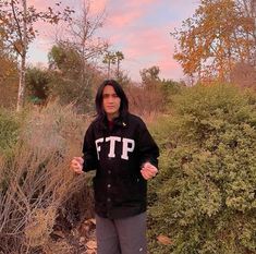 a woman standing in front of bushes with her hands out to the side, wearing a black hoodie that says ftp