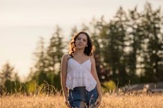 a woman standing in the middle of a field with trees in the backgroud