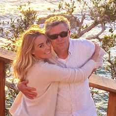 a man and woman hugging each other while standing on a wooden deck near the ocean