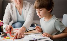 a woman sitting at a table with a little boy in front of him while he is drawing