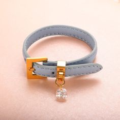 a blue leather bracelet with a gold clasp and a white diamond on the end, sitting on a pink surface