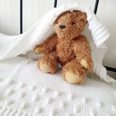 a brown teddy bear sitting under a white blanket on top of a crocheted bed