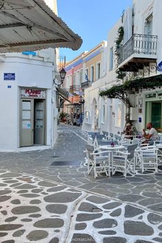 an empty street with tables and chairs on the side walk, in front of white buildings