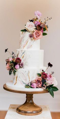 a white wedding cake with pink flowers and greenery on top, sitting on a gold pedestal