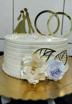 a wedding cake decorated with white flowers and gold rings on a table next to a wall