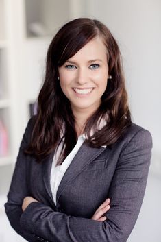 a smiling woman with her arms crossed in front of bookshelves and she is wearing a gray blazer