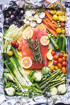 an assortment of fresh vegetables and fish on tin foil with lemons, asparagus, tomatoes, carrots, cucumbers, broccoli, radishes