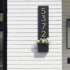 a house number sign mounted to the side of a white building with flowers growing out of it