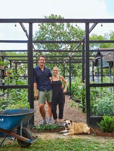 a man and woman standing next to a dog in a garden with a wheelbarrow