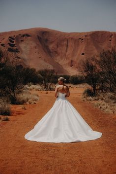 a woman in a wedding dress standing on a dirt road with mountains in the background