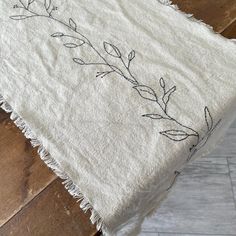 a white table cloth with black embroidery on the edge and fringed edges, sitting on top of a wooden floor