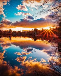 the sun is setting over a lake with trees and clouds reflected in it's water