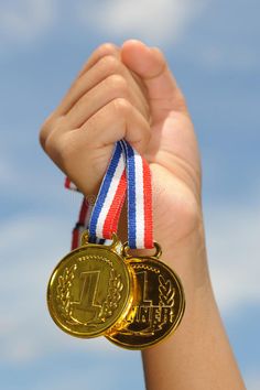 a hand holding two gold and silver medals against a blue sky with clouds in the background