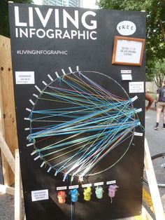 a black sign with many different colored pins on it and people walking in the background